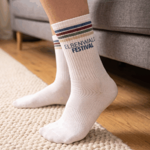 Streifen Socken - Elbenwald Festival Weiße Socken mit dem Aufdruck "Elbenwald Festival" und bunten Streifen, getragen von einer Person, die auf einem Teppich steht. Die Socken sind ein beliebtes Merchandise-Produkt für Fans des Festivals, das Musik und Nerdkultur kombiniert.
