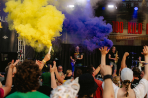 100 Kilo Herz Buntes Festivalpublikum beim Elbenwald Festival, das mit gelbem und lila Rauch von Pyrotechnik begeistert. Musiker auf der Bühne sorgen für eine lebendige Atmosphäre, während die Menge jubelt und die Hände in die Luft wirft.