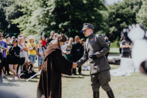 501st Legion Zwei Cosplayer, ein Jedi und ein Imperialer Offizier, schütteln sich die Hände auf dem Elbenwald Festival. Im Hintergrund applaudiert eine begeisterte Menge von Fans in verschiedenen Kostümen, umgeben von einer lebhaften Festivalatmosphäre.