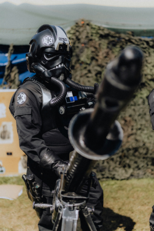 501st Legion Ein kostümierter Teilnehmer des Elbenwald Festivals trägt eine schwarze Uniform mit einem Helm im Star-Wars-Stil und hält eine große Waffe. Im Hintergrund sind militärische Elemente und eine grüne Plane sichtbar, die die Atmosphäre des Festivals unterstreichen.
