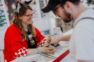 Ichiban Kuji Eine Verkäuferin mit Katzenohren lächelt, während sie einem Besucher beim Elbenwald Festival eine Box reicht. Im Hintergrund sind bunte Flyer und Merchandise-Artikel zu sehen, die die kreative und lebendige Atmosphäre des Festivals widerspiegeln.