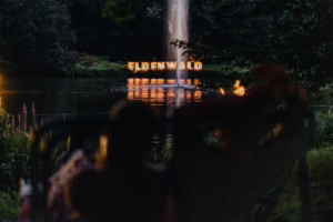 Beleuchteter Schriftzug "Elbenwald" am Ufer eines ruhigen Teiches, umgeben von Bäumen und Blumen. Im Hintergrund sprudelt ein Wasserfall, während Besucher in gemütlichen Stühlen die Atmosphäre des Festivals genießen.
