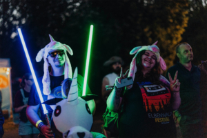 Zwei Festivalbesucher in Einhornmützen halten leuchtende Lichtschwerter und posieren fröhlich für die Kamera. Im Hintergrund sind weitere Teilnehmer des Elbenwald Festivals zu sehen, die die entspannte Atmosphäre genießen.