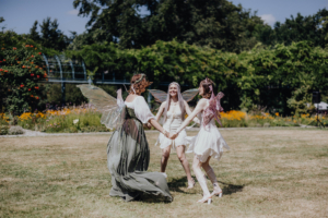 Drei Frauen in fantasievollen Feenkostümen tanzen im Kreis auf einer Wiese beim Elbenwald Festival. Bunte Blumen und eine grüne Kulisse umgeben die fröhliche Szene, die das kreative und verspielte Ambiente des Festivals widerspiegelt.