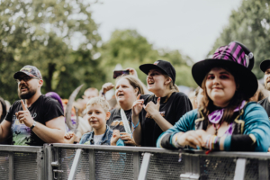 Festivalbesucher beim Elbenwald Festival jubeln und tanzen vor der Bühne. Die Menge, bestehend aus Fans unterschiedlichster Altersgruppen, zeigt Begeisterung für die Musik und das Event. Der Fokus liegt auf der lebhaften Atmosphäre und dem Gemeinschaftsgefühl.