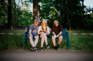 Drei Freunde sitzen auf einer Bank beim Elbenwald Festival, tragen kreative Mützen und genießen die Atmosphäre. Umgeben von Natur und Festival-Elementen strahlen sie Freude und Gemeinschaftsgefühl aus. Ideal für Fans von Musik und Nerd-Kultur.