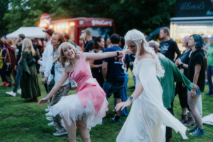 Zwei Frauen in Kostümen tanzen fröhlich auf dem Elbenwald Festival, umgeben von einer bunten Menge. Die Atmosphäre ist lebhaft, mit Musik und Freundschaft, während die Teilnehmer die einzigartige Mischung aus Musik und Nerdkultur genießen.