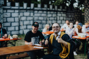 Zwei Teilnehmer des Elbenwald Festivals sitzen an einem Tisch, lächeln und geben Daumen hoch. Im Hintergrund ist eine Mauer zu sehen, während andere Besucher an weiteren Tischen sitzen. Die Atmosphäre ist entspannt und freundlich, ideal für Fans von Musik und Nerdkultur.