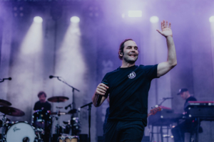 Bosse Ein Musiker auf der Bühne des Elbenwald Festivals, umgeben von bunten Lichtern und Nebel. Im Hintergrund sind Bandmitglieder an ihren Instrumenten zu sehen, während der Künstler das Publikum mit einem Lächeln und einer Handbewegung begeistert.