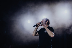 Bosse Ein Sänger steht auf der Bühne des Elbenwald Festivals, umgeben von Nebel und Bühnenlichtern. Er hält ein Mikrofon in der Hand und begeistert das Publikum mit seiner Performance. Die Atmosphäre ist energiegeladen und festlich.