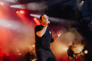Bosse Ein Sänger tritt auf der Bühne des Elbenwald Festivals auf, umgeben von bunten Lichtern und Nebel. Die Atmosphäre ist lebhaft und energiegeladen, während die Band im Hintergrund spielt. Fans genießen die Mischung aus Musik und Nerdkultur.