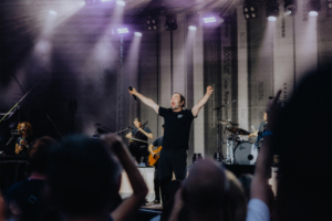 Bosse Ein Sänger begeistert das Publikum auf der Bühne des Elbenwald Festivals, umgeben von Bandmitgliedern und einer lebendigen Atmosphäre. Die Lichter strahlen, während Fans begeistert mit den Händen in der Luft feiern.