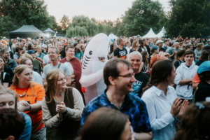 Eine lebhafte Menschenmenge beim Elbenwald Festival, mit Teilnehmern in kreativen Kostümen, darunter ein aufblasbarer Hai. Die Atmosphäre ist festlich und energiegeladen, während die Besucher Musik genießen und klatschen.