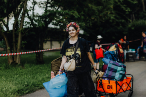Junge Frau mit Blumenkranz trägt Gepäck in einem Wagen beim Elbenwald Festival. Im Hintergrund sind weitere Festivalbesucher mit Taschen und Rucksäcken zu sehen. Die Atmosphäre ist entspannt und festlich, ideal für Fans von Musik und Nerdkultur.