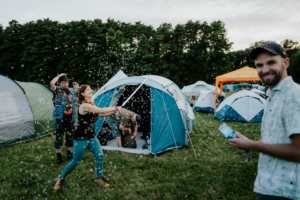 Festivalbesucher beim Elbenwald Festival, umgeben von Zelten, während sie Seifenblasen machen. Die fröhliche Atmosphäre spiegelt die Mischung aus Musik und nerdiger Kultur wider, mit Freunden, die gemeinsam Spaß haben und die Natur genießen.