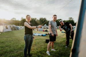 Drei junge Erwachsene auf dem Elbenwald Festival, die ein Zelt aufbauen. Die Szene zeigt eine entspannte Festivalatmosphäre mit Zelten im Hintergrund und einer warmen Abendstimmung. Ideal für Fans von Musik und Nerdkultur.