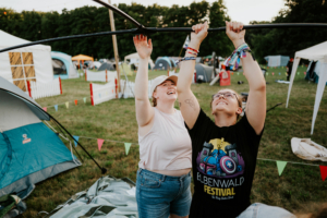 Zwei Frauen bauen gemeinsam ein Zelt auf dem Elbenwald Festival auf. Sie lachen und arbeiten zusammen, während im Hintergrund weitere Zelte und bunte Wimpel zu sehen sind. Das Festival verbindet Musik und Nerdkultur in einer lebhaften Atmosphäre.