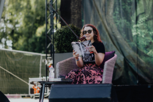 Carina Schnell Eine Rednerin sitzt auf einer Bühne beim Elbenwald Festival, während sie aus einem Buch vorliest. Sie trägt eine Sonnenbrille und ein T-Shirt mit Aufdruck. Im Hintergrund sind grüne Bäume und Festival-Ausstattung zu sehen.