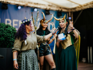 Zwei Cosplayerinnen auf der Bühne des Elbenwald Festivals, eine mit Mikrofon und einer Geschenkbox, die andere in einem Loki-Kostüm mit Hörnern. Im Hintergrund ein grüner Busch und eine festliche Atmosphäre, die die kreative Gemeinschaft feiert.