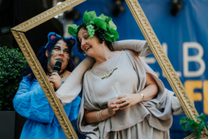 Cosplay Catwalk mit SaKiDe Zwei Frauen in fantasievollen Kostümen posieren mit einem goldenen Bilderrahmen auf dem Elbenwald Festival, umgeben von grünen Pflanzen. Die Frau links hält ein Mikrofon, während die andere fröhlich in die Kamera lächelt. Bunte und kreative Atmosphäre des Festivals.
