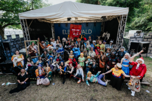 Cosplay Catwalk mit SaKiDe Gruppenshot von Teilnehmern des Elbenwald Festivals, die in bunten Kostümen aus verschiedenen Fantasiewelten posieren. Die Bühne im Hintergrund zeigt das Festival-Logo, während die fröhlichen Besucher eine lebendige Atmosphäre schaffen.