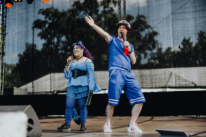 Elbenwald Cosplay Contest Zwei Moderatoren auf der Bühne des Elbenwald Festivals, gekleidet in fantasievolle Kostüme. Die Frau trägt ein hellblaues Outfit mit lila Haaren, der Mann ein sportliches blaues Ensemble. Beide halten Mikrofone und interagieren mit dem Publikum.