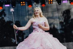 Elbenwald Cosplay Contest Eine lächelnde Frau in einem glitzernden, rosa Kleid und einer Krone steht auf einer Bühne beim Elbenwald Festival. Sie hält einen Zauberstab und strahlt Freude aus, während die bunten Lichter im Hintergrund leuchten.