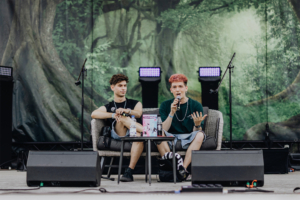 darkviktory & Kostas Kind Zwei junge Männer sitzen auf einer Bühne beim Elbenwald Festival und diskutieren. Der Hintergrund zeigt eine grüne, mystische Waldlandschaft. Auf dem Tisch befindet sich ein Magazin, während Mikrofone für die Veranstaltung bereitstehen.