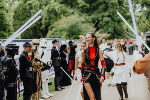 Teilnehmer des Elbenwald Festivals in Kostümen, darunter Stormtrooper und Jedi, laufen mit Lichtschwertern durch eine grüne Parklandschaft. Die Atmosphäre ist lebhaft und kreativ, was die Verbindung von Musik und Nerdkultur widerspiegelt.