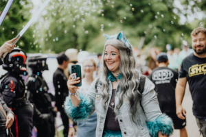 Festivalbesucher mit grauen Haaren und bunten Accessoires, die ein Smartphone in der Hand hält, umgeben von schaumigen Blasen und Cosplayern, während sie beim Elbenwald Festival Spaß hat. Hintergrund mit Menschen in Kostümen und lebendiger Festivalatmosphäre.