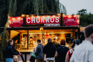 Churros-Stand auf dem Elbenwald Festival, mit Besuchern, die sich für leckere Snacks und Getränke entscheiden. Die beleuchtete Verkaufsbude bietet Churros, Softeis und Kaffee in einer lebhaften Festivalatmosphäre.