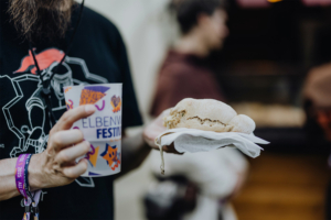 Ein Besucher des Elbenwald Festivals hält ein Getränk und ein belegtes Brötchen in der Hand. Im Hintergrund sind andere Festivalteilnehmer zu sehen, die die Atmosphäre des Events genießen.
