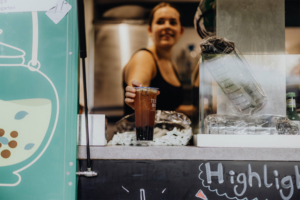 Eine lächelnde Verkäuferin reicht ein Getränk am Elbenwald Festival, das eine Mischung aus Musik und Nerd-Kultur bietet. Der Kiosk ist mit bunten Grafiken gestaltet, und im Hintergrund sind weitere Getränke sichtbar.