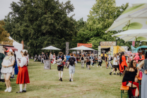 Besucher schlendern über das Gelände des Elbenwald Festivals, umgeben von bunten Ständen und Essensständen. Die Atmosphäre ist lebhaft und einladend, während Fans von Musik und Popkultur in Kostümen feiern und das Festival genießen.