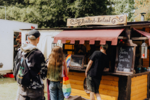 Food Besucher stehen am Stand "Der Handbrot-Heiland" auf dem Elbenwald Festival und betrachten das Menü mit verschiedenen Handbrot-Spezialitäten wie Käse-Schinken und Käse-Spinat-Feta. Bunte Taschen und eine entspannte Atmosphäre prägen die Szene.
