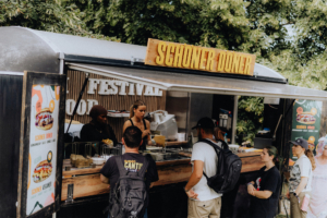 Food Ein Foodtruck mit dem Namen "Schöner Döner" bietet Besuchern des Elbenwald Festivals köstliche Snacks an. Im Vordergrund stehen mehrere Personen in Warteschlange, während die Verkäuferinnen die Bestellungen entgegennehmen.