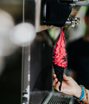 Food Ein Hand hält ein schwarzes Waffelhorn mit leuchtend rotem Softeis, das aus einer Maschine herausgespritzt wird. Der Hintergrund zeigt verschwommene Festivalatmosphäre, passend zum Elbenwald Festival, wo Genuss und Spaß im Vordergrund stehen.