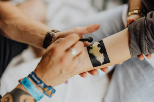 Glitzer-Tattoos Ein Festivalbesucher erhält ein Glitzer-Tattoo auf dem Elbenwald Festival. Die Person trägt ein schwarzes T-Shirt und bunte Festival-Armbänder, während ein Künstler das Tattoo mit einem Pinsel gestaltet. Die Atmosphäre ist kreativ und lebhaft.