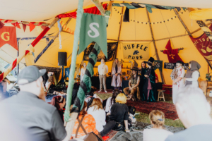 MMM Larp Bunte Zeltatmosphäre beim Elbenwald Festival, mit Teilnehmern in Kostümen, die eine Szene aufführen. Im Hintergrund sind Flaggen von Hogwarts-Häusern und eine Bühne sichtbar, während das Publikum gespannt zusieht.