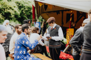 MMM Larp Besucher des Elbenwald Festivals sitzen an einem Holztisch, während ein verkleideter Händler mit einem Besucher spricht. Im Hintergrund sind bunte Wimpel und weitere Festivalteilnehmer zu sehen, die die lebendige Atmosphäre des Events unterstreichen.