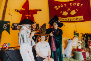 MMM Larp Eine Frau sitzt mit einem Zauberer-Hut auf dem Kopf, während zwei verkleidete Personen um sie herum stehen. Im Hintergrund sind Flaggen und Dekorationen des Elbenwald Festivals zu sehen, die eine magische Atmosphäre schaffen.
