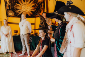 MMM Larp Ein Mädchen sitzt mit einem Zauberhut auf dem Kopf, während eine andere Person den Hut anpasst. Im Hintergrund sind weitere Teilnehmer in Kostümen zu sehen, umgeben von dekorativen Elementen im Zelt während des Elbenwald Festivals.