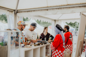 Ichiban Kuji Lotterie Besucher des Elbenwald Festivals stehen an einem Stand, während sie Merchandise und Fanartikel durchstöbern. Die Atmosphäre ist lebhaft und bunt, mit einer Mischung aus Cosplay und Festivalstimmung unter dem Zelt.