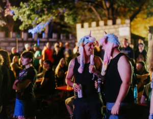 Zwei Männer in bunten Einhornmützen lachen und unterhalten sich auf dem Elbenwald Festival, umgeben von einer fröhlichen Menge. Die Atmosphäre ist lebhaft und zeigt die Kombination aus Musik und nerdiger Kultur.