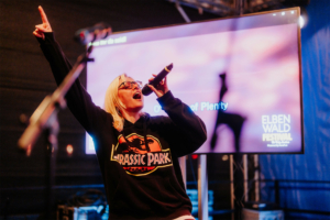 Eine Sängerin in einem Jurassic Park-Hoodie begeistert das Publikum beim Elbenwald Festival. Sie hält ein Mikrofon und singt leidenschaftlich, während im Hintergrund eine Projektionsleinwand mit dem Festival-Logo und dem Titel „Order of Plenty“ zu sehen ist.
