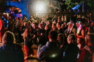 Menschenmenge beim Elbenwald Festival, die auf einer Bühne tanzt und feiert, umgeben von bunten Lichtern und kreativen Kostümen. Die Atmosphäre ist lebhaft und freundlich, während sich Teilnehmer in einem einladenden, nerdigen Ambiente versammeln.