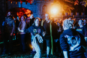 Festivalbesucher tanzen begeistert auf dem Elbenwald Festival, umgeben von bunten Lichtern und einer festlichen Atmosphäre. Die Teilnehmer tragen kreative Outfits und genießen die Mischung aus Musik und Gemeinschaft.