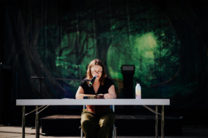 Lesung Karolyn Ciseau Eine Frau mit Brille sitzt an einem Tisch und liest in ein Mikrofon, während sie auf der Bühne des Elbenwald Festivals steht. Im Hintergrund ist eine grüne Waldlandschaft zu sehen, die die Atmosphäre des Festivals unterstreicht.