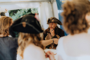 Meet and Greet Katerfahrt Ein Pirat im Kostüm sitzt am Tisch und betrachtet die Besucher des Elbenwald Festivals, während mehrere Personen mit Hüten ihm aufmerksam zuhören. Die Szene vermittelt die lebendige Atmosphäre des Festivals, das Musik und Nerdkultur vereint.