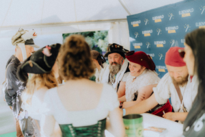 Meet and Greet Katerfahrt Besucher in Kostümen interagieren mit Darstellern am Elbenwald Festival. Die Szene zeigt eine lebhafte Atmosphäre mit Piraten und Fantasiekostümen, während die Teilnehmer Autogramme sammeln und sich austauschen.